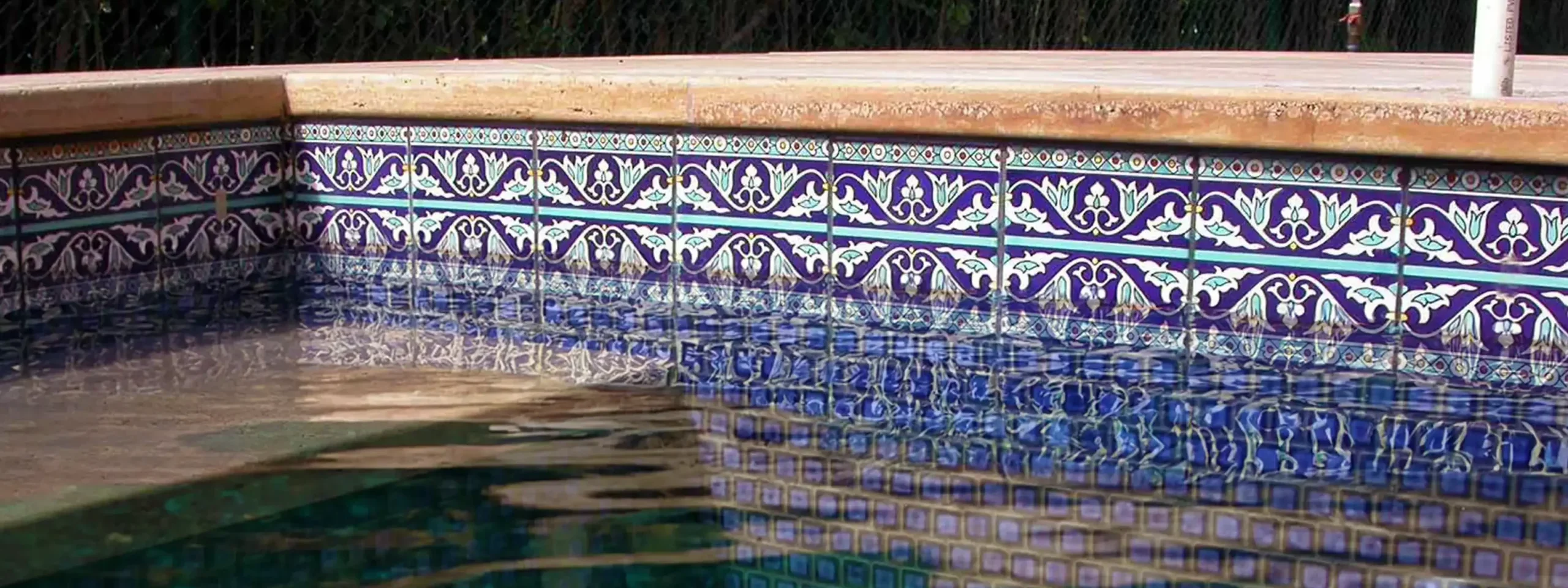 The corner of a swimming pool featuring a waterline decorated with hand-decorated ceramic tiles with a repeating blue, white, and purple floral and geometric pattern. The pool water is clear and reflects the pattern of the tiles, which continue onto a step within the pool. The pool deck is made of light tan stone.