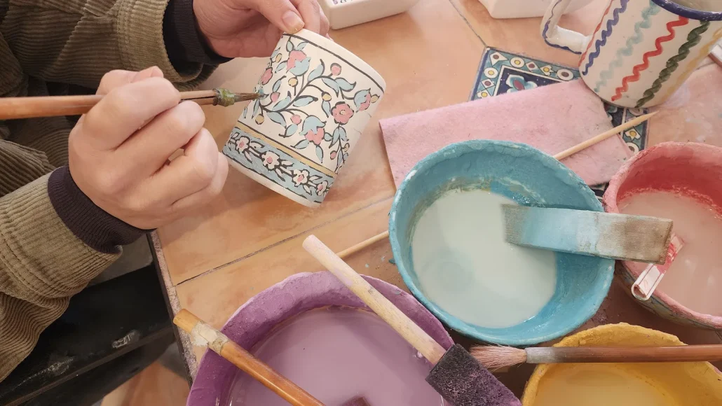 A high-angle close-up photograph captures a person's hands carefully painting a white ceramic mug with blue and pink floral patterns using a thin brush and light blue glaze. The mug is being held over a tiled wooden table covered with various bowls of colorful ceramic glazes, including light blue, purple, pink, and yellow, along with several wooden brushes and sponges. In the background, another finished ceramic mug with green and red patterns is visible.