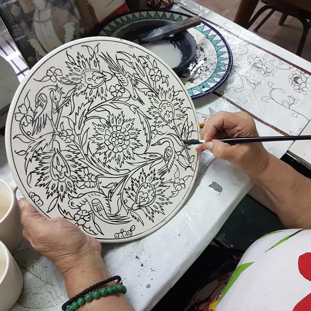 A close-up photograph shows a pair of older hands, one wearing a green bead bracelet, using a fine brush to carefully hand paint an intricate black-line floral and bird design onto a round ceramic plate. The plate is being decorated in the studio of Balian Pottery of Jerusalem.